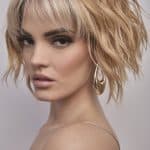 Model wearing a blonde, textured, layered wig with bangs and tousled style.